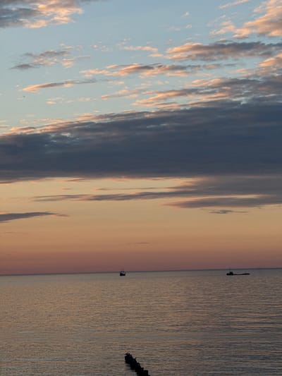 A serene seascape at dusk captures the horizon over the Baltic Sea in Ustka, Poland. The sky transitions from a soft blue at the top to warm orange and pink hues near the water, painted with scattered clouds tinged with pink and gray. Two vessels, one a sailing ship and the other a modern freighter, are visible as dark silhouettes on the calm, rippling water. In the foreground, a series of wooden pilings, partially submerged, break the surface of the sea.