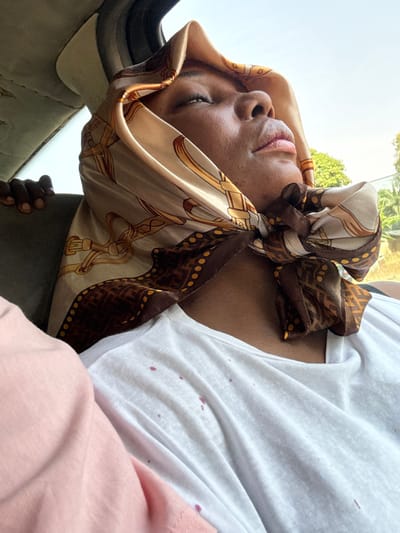 The image depicts an interior scene, likely within a vehicle, during daylight hours, in the city of Mayo, Nigeria. A dark-skinned individual, seen from the mid-chest upward, is the primary subject. Their head is tilted slightly upward, and their eyes are closed or gazing skyward. They wear a light-toned silk headscarf patterned with gold belts and chains, featuring a dark brown border with yellow dots and a repeating 'F' design, tied under the chin. Below the scarf, a white crew-neck t-shirt is visible, marked with multiple scattered reddish-purple stains across the chest area. A pink fabric is partially visible on the immediate left. In the upper left background, the partial hand of another dark-skinned individual is visible. The vehicle's interior headliner, appearing worn, is visible above the hand. Through a window on the right, bright daylight, green foliage, and indistinct background elements are observable.