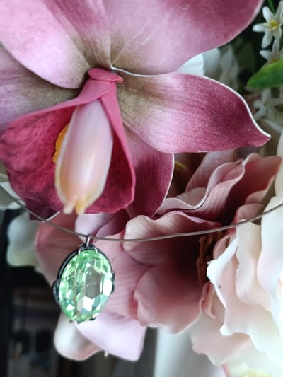 A close-up, eye-level, outdoor shot features a delicate silver necklace with a prominent, oval, faceted light green gemstone pendant hanging in the foreground. The pendant is the primary subject, set against a blurred backdrop of artificial pink and white flowers. The necklace rests on what appears to be a wire. The gemstone is the only clearly focused object in the frame. The background flowers are soft and out of focus, with shades of deep pink, burgundy, and pale pink, creating a romantic and slightly ethereal atmosphere. The lighting is soft, highlighting the facets of the gemstone and the textures of the petals. There is no indication of the city of Évreux, France, or any specific environmental context.