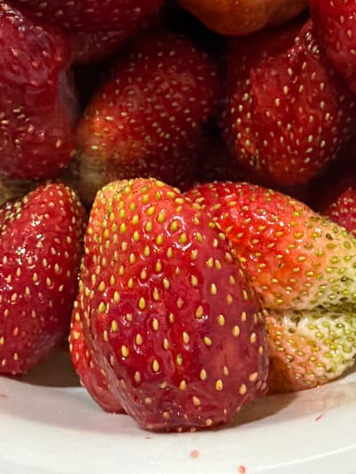 This is a close-up, high-angle shot of a pile of fresh strawberries, likely in a bowl or container, given the white edge visible on the right side of the frame. The strawberries are predominantly a vibrant red, with some showing greener tips, suggesting they are in various stages of ripeness. Each strawberry is covered in numerous tiny yellow seeds, characteristic of the fruit. The surface of the strawberries appears slightly wet and shiny, hinting at them being recently washed or juicy. The lighting is bright, emphasizing the rich colors and textures of the fruit. No people or text are visible in the image. The context provided indicates the location is Шемордан, Russia, but the image itself offers no specific visual cues to confirm this. The scene is entirely focused on the strawberries, presenting them as the sole subject.