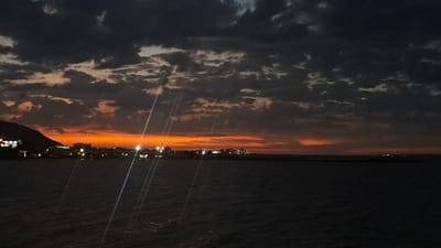 Night seascape features a skyline of coastal buildings, faintly illuminated against a fiery orange horizon and dark, cloudy sky. The dark water reflects the sky, punctuated by vertical light streaks from an unknown source. A distant landmass is visible at the left. The scene is dominated by shades of orange, black, and gray, emphasizing the transition from day to night.