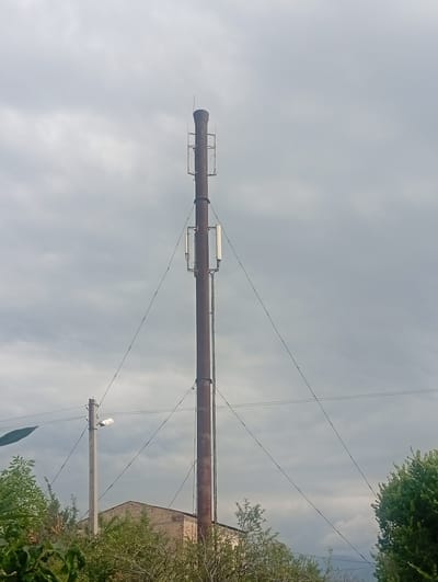 In the city of Arashan, Kyrgyzstan, a tall, rusty smokestack stands against a cloudy sky. Mounted on the smokestack are three rectangular antennas, two on the upper section and one lower down, suggesting it may be repurposed as a cell tower. Wires are strung from the smokestack to a concrete utility pole visible to the left, which supports a streetlamp. In the foreground, lush green trees and foliage partially obscure a low-rise brick building. The scene is set outdoors, under a heavily overcast sky, indicating potentially inclement weather.