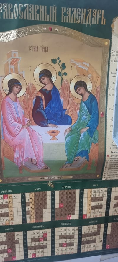 This is an image of a religious calendar featuring an icon of the Holy Trinity. The icon depicts three angels seated around a table, painted in traditional Orthodox style with halos. The calendar itself is oriented vertically, with text in Russian. The main title at the top reads "ПРАВОСЛАВНЫЙ КАЛЕНДАРЬ" (Orthodox Calendar). Below the icon, the text "ИКОНА «СВЯТАЯ ТРОИЦА»" (Icon "Holy Trinity") is visible.
The calendar portion shows months with dates and days of the week represented by abbreviations. Months such as ФЕВРАЛЬ (February), МАРТ (March), АПРЕЛЬ (April), МАЙ (May), АВГУСТ (August), СЕНТЯБРЬ (September), ОКТЯБРЬ (October), and НОЯБРЬ (November) are visible. There are also some specific dates marked with red, possibly indicating holidays or important religious events.
The overall aesthetic is religious and traditional. The image appears to be a printed calendar, likely for personal use in a home or religious institution. The color palette is rich with gold, blues, pinks, and greens. The lighting is consistent and suggests an indoor setting. There are no explicit indications of time of day, weather, or specific emotions conveyed by the figures in the icon, which are depicted in a serene and symbolic manner. The calendar is the primary functional object, and the icon serves as its central decorative and spiritual element.