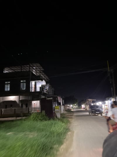 The image depicts a nighttime street scene in the city of Tanjungkarang, Indonesia. A multi-story building with illuminated windows and a metal roof structure stands on the left side of the frame, bordered by a patch of tall grass. The street itself is visible on the right, with a dark-colored SUV driving away from the camera. Along the street, there are several small shops or stalls with lights illuminating their fronts. A person is partially visible in the foreground on the right, suggesting the photo was taken from a moving vehicle, possibly a motorcycle. The sky is dark, indicating it is nighttime, and power lines are visible overhead.