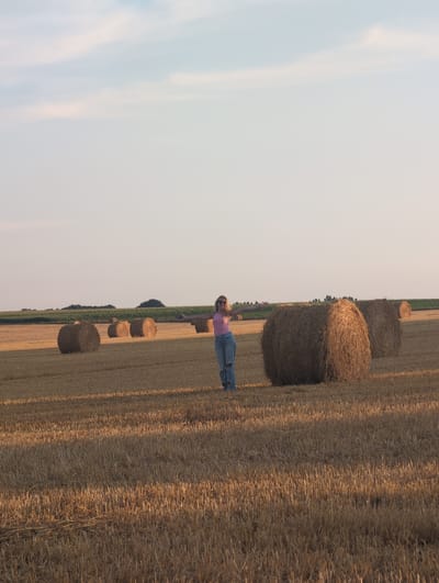 An outdoor, medium-shot photograph captures a woman standing in a harvested field with round hay bales scattered across the landscape. The woman, positioned slightly to the left of the frame's center, wears a pink tank top and ripped blue jeans. She is facing the camera with her arms outstretched to the sides and appears to be smiling, with sunglasses on her face. Behind her, several large, round hay bales are visible, most notably a prominent one to her right. The field is covered in dry, golden straw, with stubble from the harvested crop. In the background, a strip of green vegetation, possibly a field of corn or trees, separates the harvested field from the pale blue sky. The lighting suggests it is late afternoon or early evening, casting long shadows across the field. The location is identified as Alvimare, France.