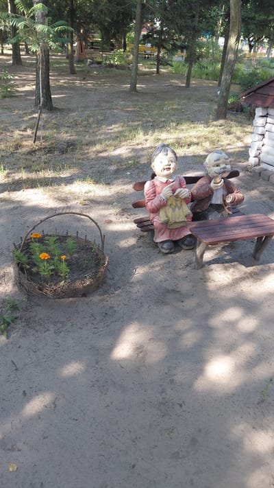 In Borodianka, Ukraine, on a sandy ground with scattered dry grass and tree shadows, a woven basket containing several small orange flowers sits to the left of the frame. To the right, two garden gnomes are seated on a wooden bench. The gnome on the left, a female figure wearing a pink dress and black shoes, is depicted knitting a yellow garment. The gnome on the right, a male figure with a mustache, wears a brown jacket and holds a pipe to his mouth. A small wooden table is positioned in front of the bench. In the background, trees and a wooden structure are visible.