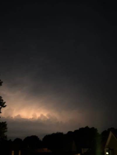 The image depicts a nighttime scene with a dark sky illuminated by a distant lightning strike or a similar light source behind a dense line of trees. The foreground shows silhouetted trees and what appear to be the roofs of buildings. The location is the city of Putten, Netherlands. The overall atmosphere is one of a storm at night.