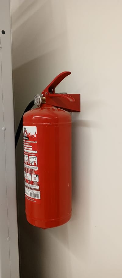 A red fire extinguisher is mounted on a light-colored wall. The fire extinguisher is a powder type and has a label with Cyrillic text and pictograms indicating its use. It is attached to a red bracket. To the left of the fire extinguisher is a portion of a light-colored vertical structure, possibly a cabinet or pillar, with a dark circular element visible on it. The wall exhibits some shadow patterns, suggesting illumination from an unseen source. The environment appears to be indoors, possibly in an office or public building. There are no people visible in the image.
