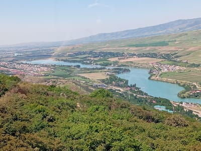 An aerial view of the city of Chinarkent, Uzbekistan, reveals a landscape dotted with urban and natural features. The topography includes undulating hills, with the foreground dominated by verdant vegetation. A winding body of water, likely a river or lake, cuts through the landscape, its turquoise color contrasting with the surrounding greenery. Buildings and infrastructure are interspersed across the terrain, suggesting a blend of residential and commercial development. In the distance, a range of mountains provides a backdrop, adding depth to the vista. The sky is hazy, casting a soft, diffused light over the scene.