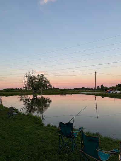 The image depicts a serene natural scene, likely at dusk, given the soft, pastel colors in the sky that transition from a light blue to shades of pink and orange. The setting appears to be a body of water, possibly a lake or river, reflecting the sky and the surrounding landscape.
In the foreground, two camping chairs are set up on a grassy bank, suggesting a leisurely activity like fishing or simply enjoying the view. One chair is green with orange trim, and another, partially visible, appears to be a similar style. Fishing rods are also visible, propped up near the water's edge, indicating that fishing is taking place or has recently occurred.
Across the water, a line of trees forms the horizon, with a solitary, large tree with bare branches standing prominently in the mid-ground, its reflection clearly visible in the still water. Power lines stretch across the sky, a man-made element contrasting with the natural beauty. In the distance, across the water, some tents and a white vehicle are parked on the grassy bank, suggesting a campsite.
The overall mood is peaceful and tranquil, evoking a sense of calm and relaxation associated with spending time outdoors at sunset. There is no visible text in the image. The location context provided is Kommunar, Russia.