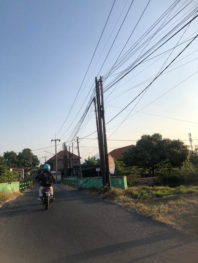 A person rides a motorcycle down a paved road in Kalidawir, Indonesia. The rider is wearing a blue helmet and a dark jacket. The road is bordered by green metal railings and dry grass. In the background, there are several buildings with red tile roofs, lush green trees, and a clear blue sky. Numerous power lines crisscross the sky, supported by wooden poles. The scene is bathed in the warm light of what appears to be either early morning or late afternoon.