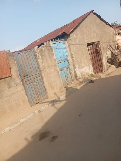 This is a medium-angle, full-shot photograph of a street scene in Gwagwa, Nigeria. The sky is clear and blue. In the center-left of the frame, a light-colored cinder block wall with a rusty, blue metal gate is visible. The gate is partially open and appears to be two panels. To the right of this gate, a blue metal door is visible. Further to the right, a building with a textured, beige-colored exterior and a corrugated metal roof is present. This building has a brown, vertical metal door. In front of the building, there is a slight rise in the ground covered in the same textured material. To the far right, a portion of a large, rusty metal container or tank is visible. On the ground in front of the building, a single dark-colored sandal is lying. The ground is a dirt road with some gravel. There are two dark wet patches on the road in the foreground. Shadows are cast across the road from an unseen source, suggesting it is daytime.