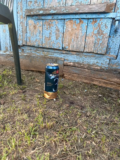 A single, unopened can of beer, specifically "Battle #3" brand, sits upright on dry grass and dirt. The can is primarily dark green with gold and white lettering, featuring a large "3" on its side. Behind the can, a weathered, light blue wooden structure with peeling paint and visible grain dominates the background. A dark green chair leg is visible on the left side of the frame. The overall environment appears to be outdoors, possibly in a rural or neglected urban setting. The location is identified as Chuya-Ataevo, Russia.