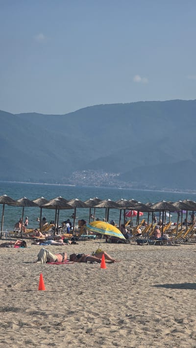 The image captures a sunny daytime scene on a sandy and pebbly beach in Asprovalta, Greece. The sky is a light, clear blue with a few small, faint white clouds. In the background, a range of dark blue-green mountains spans the horizon across a calm body of water, which is a deep blue hue. A settlement or town with numerous buildings is visible on the lower slopes of these distant mountains.
The sea contains several individuals swimming and standing in the shallows. Along the water's edge, a continuous row of straw-thatched beach umbrellas provides shade for an array of yellow sun loungers, many of which are occupied by people relaxing.
The sandy and pebbly beach occupies the foreground and mid-ground. Several individuals are sunbathing directly on the sand. One person is lying on a red mat under a yellow and blue striped beach umbrella. Another person lies on the sand with a light-colored fabric covering their lower body. Two orange traffic cones are positioned in the sand in the foreground; one is closer to the viewer, and the second is placed beside a person sunbathing.