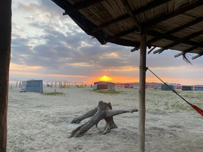 The image captures a sunset view from under a thatched roof structure on the beach of General Villamil Playas, Ecuador. The sun is setting on the horizon, partially obscured by clouds. The beach is sandy with scattered patches of grass and driftwood. Several small structures or beach huts are visible in the distance, along with beach furniture like chairs and umbrellas. A large piece of driftwood resembling a tree stump sits prominently in the foreground. To the left and right, wooden posts frame the view, suggesting the perspective is from within a sheltered area. A hammock is partially visible on the right side of the frame.