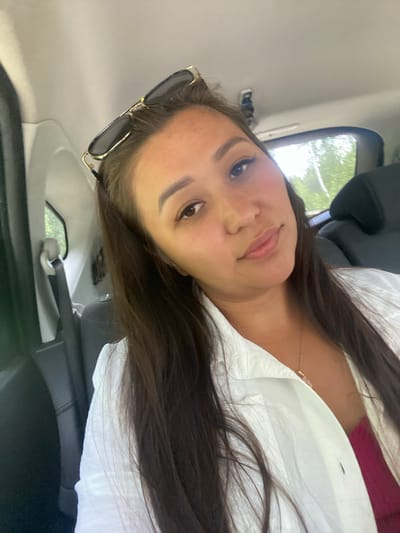 A young woman with long dark hair, wearing sunglasses on her head and a white open shirt over a pink top, is taking a selfie in the back seat of a car. The interior of the car is visible, with black seats and a seatbelt. Outside the window, trees and greenery can be seen. The scene appears to be in a rural or natural setting, possibly on a road trip. The image was taken on a sunny day, casting shadows and highlights. The location is identified as Kashkarovo, Russia.