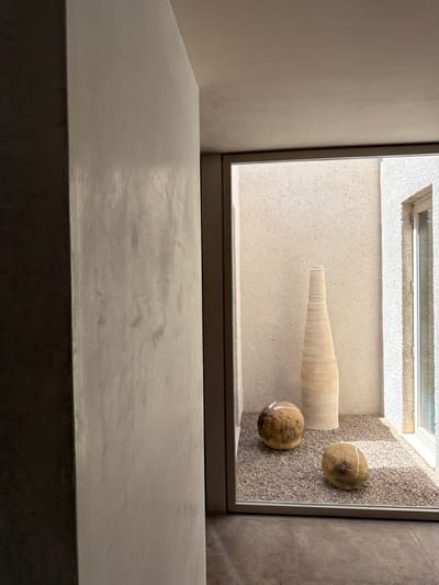 The image shows an interior courtyard with a gravel floor. Two round, textured decorative orbs with white bands are placed on the gravel. Behind the orbs, a tall, ribbed, vase-like object stands upright. The courtyard is enclosed by textured walls and a large window. The lighting suggests it is daytime. The scene appears to be located in Artà, Spain.