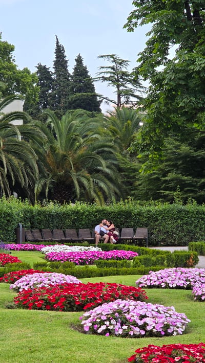 In Riva del Garda, Italy, a couple is shown in an intimate embrace on a park bench. The man, wearing a white shirt, is seated while the woman, dressed in a burgundy dress, is leaning into him. They are surrounded by lush greenery, including tall palm trees and manicured hedges. In the foreground, vibrant rows of red, pink, and white impatiens flowers are beautifully arranged on manicured green grass. The scene suggests a warm, pleasant day, likely late spring or summer, given the blooming flowers and attire of the couple. The background features more dense foliage and trees, with a bright, overcast sky visible above. The overall atmosphere is romantic and serene, capturing a moment of connection amidst a picturesque garden setting. No visible text is present in the image.