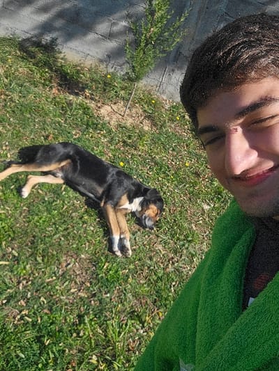 A selfie-style photograph depicts a young male and a medium-sized dog outdoors in Buriani, Georgia, under direct sunlight. The male subject occupies the right foreground, presenting a smiling expression with dark hair, partially closed eyes, and a visible scab on the bridge of his nose. He is clad in a bright green, textured garment. The dog, predominantly black with tan markings on its legs, chest, and face, and white paws, lies on its left side on green grass in the mid-left portion of the frame, appearing to be asleep or resting with its eyes closed. The grassy ground contains scattered small yellow flowers and dry patches. A concrete block wall forms the background, partially shaded, with a slender green sapling growing near it. Shadows are distinctly cast across the scene.