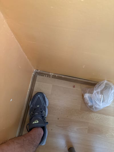 The image displays an indoor corner, looking down at a light tan-colored wall and a light wooden floor. A dark brown transition strip runs along the base of the wall where it meets the floor, with some light-colored residue, possibly caulk, visible.

In the bottom right, a person's lower leg, covered in hair, is visible, wearing a dark grey New Balance 9060 sneaker with yellow accents. The New Balance brand name and "9060" model number are clearly visible on the shoe's tongue.

On the wooden floor, near the wall in the upper right, rests a clear plastic bag containing some white or light-colored items, possibly a small cloth or tissue. Faint, handwritten text resembling "6h" is barely discernible on a label inside the bag. To the far right, a dark grey cylindrical object, likely a furniture leg, is partially visible. The wall has some minor scuff marks and textural variations, while the wooden floor shows distinct grain patterns. The scene depicts a casual interior setting.