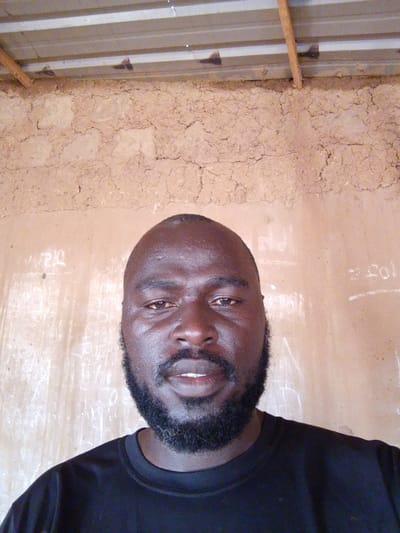 A medium, eye-level shot of a dark-skinned man with a shaved head and a full black beard. He is wearing a black shirt. Behind him is an interior wall constructed of mud brick. Above the mud brick is a corrugated metal roof supported by wooden beams. Writing is visible on the wall. The location is likely Mulke Barikin Sayejo, Nigeria.