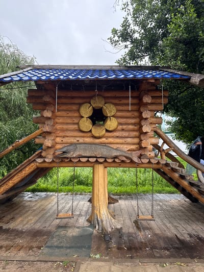 A wooden structure resembling a small log cabin with a blue tiled roof is situated outdoors. Attached to the structure are two swings, each with a wooden seat suspended by chains. A carved wooden fish is mounted horizontally on the front of the cabin. The ground beneath the structure is made of wooden planks, some of which appear wet. Surrounding the wooden structure is lush green vegetation and trees, suggesting an outdoor environment, possibly a park or recreational area. The sky is overcast.