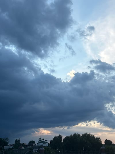 The image shows a sky with dramatic, dark clouds dominating the upper portion of the frame. A bright patch of sky, possibly illuminated by the sun, breaks through the clouds in the upper right quadrant, with lighter, wispy clouds visible there as well. Below the heavy cloud cover, the scene transitions to a glimpse of a village or town. Visible are the tops of several buildings, including what appears to be a church with multiple green domes. Trees are scattered throughout the lower part of the image, some appearing dense and dark, while others are lighter, suggesting illumination from the sky. A fence is visible along the bottom edge of the frame. The overall impression is one of an impending storm or a post-storm sky, with the village nestled beneath the dramatic celestial display. The location is identified as Zhukhavitski Sel'ski Savet, Belarus.