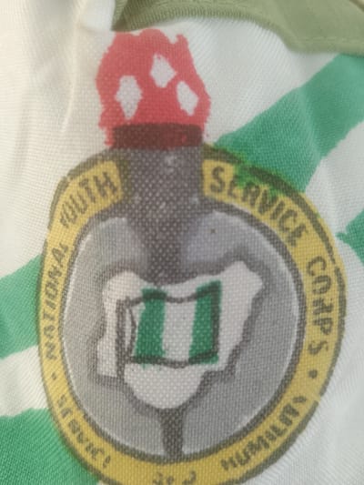 A close-up, overhead view shows a circular emblem embroidered on a white and green striped fabric. The emblem features a torch with a red and white top and a grey shaft. Encircling the torch is a yellow band with text. The words "NATIONAL YOUTH SERVICE CORPS" are visible on the upper half of the band, and "HUMILITY" is on the lower half. Within the yellow band, the emblem contains a grey circle with a depiction of Nigeria, featuring a green and white flag in the center. The fabric itself appears to be part of a garment or textile, likely a shirt or banner, with green stripes running diagonally. The image does not provide any information about the city of Majemu, Nigeria, or any specific location.