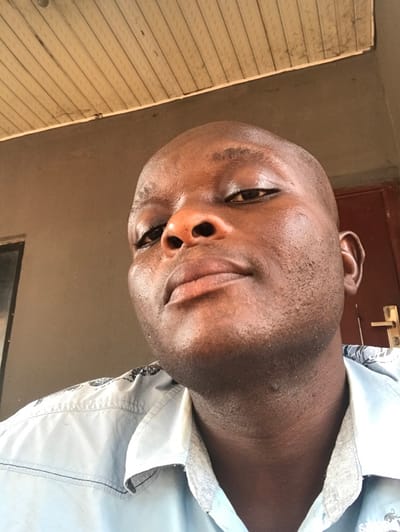 A close-up, slightly upward-angled portrait depicts an adult male of dark complexion. He has a bald head, dark eyes, a prominent nose, and full lips, maintaining a direct gaze towards the camera. He is wearing a light blue collared shirt featuring a darker, possibly patterned, design across the shoulders and upper chest. The background comprises a light-colored, slatted ceiling overhead, a plain light brown wall to the left, and a dark brown wooden door with a gold-toned handle visible on the right. A dark door frame is partially discernible. The setting is consistent with an indoor or sheltered environment. The specific location in Egboje, Nigeria, as provided, is not verifiable through visual analysis of the image content.