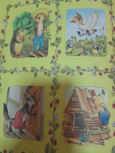 The image depicts a page from a children's book, featuring four illustrated vignettes on a light yellow background adorned with a repeating pattern of strawberries and white flowers.

The top left vignette shows a hedgehog and a rabbit wearing blue overalls and a straw hat, standing near a large head of cabbage. The rabbit appears to be speaking or singing.

The top right vignette illustrates a duck in mid-flight above a bush with two small blue and yellow birds perched on it.

The bottom left vignette features a wolf, dressed in a red apron, peeking around a wooden door.

The bottom right vignette shows a pig wearing blue overalls, building a wooden structure with planks and logs. Tools, including a saw and a toolbox, are visible near the pig.

The provided image does not contain any information or visual elements that would allow for the identification of its location in Baan Mae Nam, Thailand.