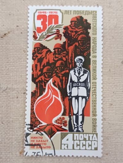 This is a postage stamp commemorating the 30th anniversary of Victory in the Great Patriotic War, from 1945 to 1975. The stamp features a depiction of Soviet soldiers, a uniformed guard, a burning eternal flame, and floral arrangements. The text on the stamp is in Russian and reads "1945-1975 30 ЛЕТ ПОБЕДЫ СОВЕТСКОГО НАРОДА В ВЕЛИКОЙ ОТЕЧЕСТВЕННОЙ ВОЙНЕ" which translates to "1945-1975 30 years of victory of the Soviet people in the Great Patriotic War." Below this, in a separate section, is text that reads "НИКТО НЕ ЗАБЫТ И НИЧТО НЕ ЗАБЫТО" meaning "No one is forgotten, and nothing is forgotten." In the bottom right corner, it says "4 ПОЧТА СССР" which means "4 Postage USSR." The stamp itself is bordered by a serrated edge characteristic of perforated stamps. The background of the image is a textured, light brown surface, likely a table or cloth. There is no identifiable context for the city of Onaldy, Turkmenistan within this image; it is a postage stamp of the Soviet Union.