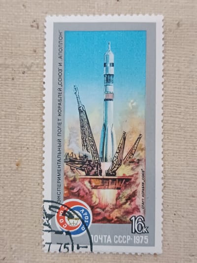 This is a postage stamp depicting a Soyuz spacecraft launching. The stamp shows the rocket on its launchpad with flames and smoke billowing from its base. Two large cranes are positioned on either side of the rocket. The background is a bright blue sky. Text on the stamp in Russian reads "ЭКСПЕРИМЕНТАЛЬНЫЙ ПОЛЕТ КОРАБЛЕЙ „СОЮЗ" И АПОЛЛОН" (Experimental flight of Soyuz and Apollo spacecraft), "СТАРТ КОРАБЛЯ „СОЮЗ" (Launch of Soyuz spacecraft), "ПОЧТА СССР-1975" (USSR Post-1975), and the denomination "16k". There is also a circular emblem with the word "APOLLO" and a globe. The stamp is postmarked with a partial cancellation.