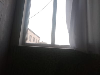 An interior view of a window in Vahdat, Tajikistan, looks out onto a street scene. The window frame is white, and a light grey curtain is visible on the right side of the image. Outside, a light-colored building with multiple windows is seen. The sky is overcast, and a wire stretches across the frame. The foreground shows a dark-colored wall with a textured pattern. No people are visible in the image.