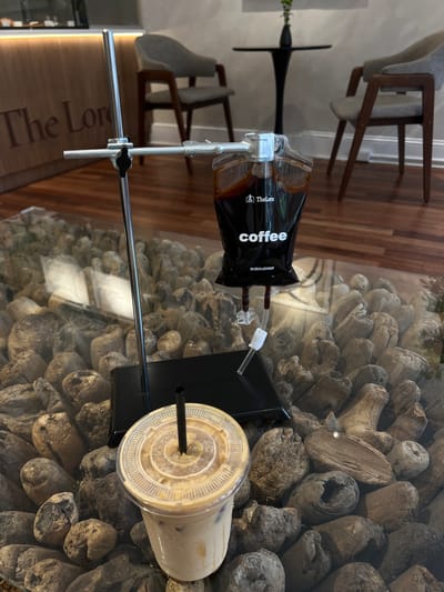 In Jenkintown, United States of America, a coffee shop scene unfolds, featuring a drip coffee bag suspended from a metal stand, with a clear plastic cup of iced coffee in the foreground. The drip bag, labeled "TheLore coffee," contains dark coffee and is connected to a tube that presumably dispenses the coffee. The iced coffee cup, topped with a clear lid and a black straw, appears to be a latte or a similar milky coffee drink with ice. Both are placed on a glass tabletop that reveals a decorative base of stones and driftwood. In the background, wooden paneling with the text "The Lore" is visible, along with chairs and a small round table. The overall setting suggests a modern and aesthetically curated coffee shop environment.