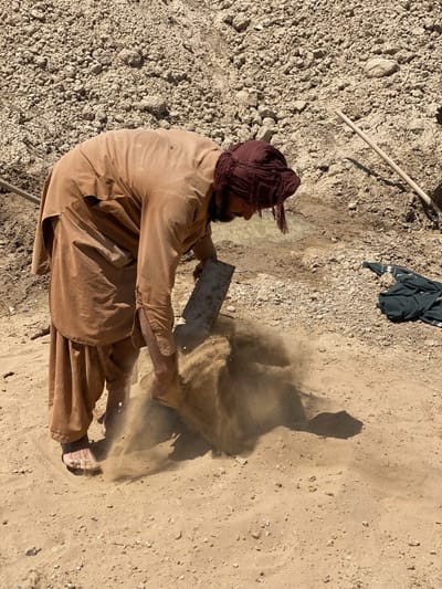 A man wearing a brown tunic and pants, a maroon head covering, and bare feet is bent over at the waist. He is sifting sand with a rectangular metal tool. Sand is being dispersed from the action of sifting. His shadow is cast onto the ground. He is on a patch of bare ground in Deh-E Bagh, Afghanistan. Behind him is a raised bank of rocky dirt. A spade lies propped against the raised bank. A piece of dark fabric lies on the ground to the right.