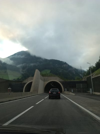 The image captures a view from a vehicle driving on a highway approaching a mountain tunnel in Rothenwand, Austria. The sky is overcast with clouds, and the surrounding mountains are covered in dense green forests, with some areas shrouded in mist. The highway has two lanes in the direction of travel, with white lane markings. In front of the vehicle, a dark-colored SUV is driving towards the tunnel entrance. The tunnel entrance is a large, arched opening in the mountainside, with concrete structures surrounding it. To the right of the road, a street lamp is visible. The overall scene suggests a cloudy and possibly damp day in a mountainous region.