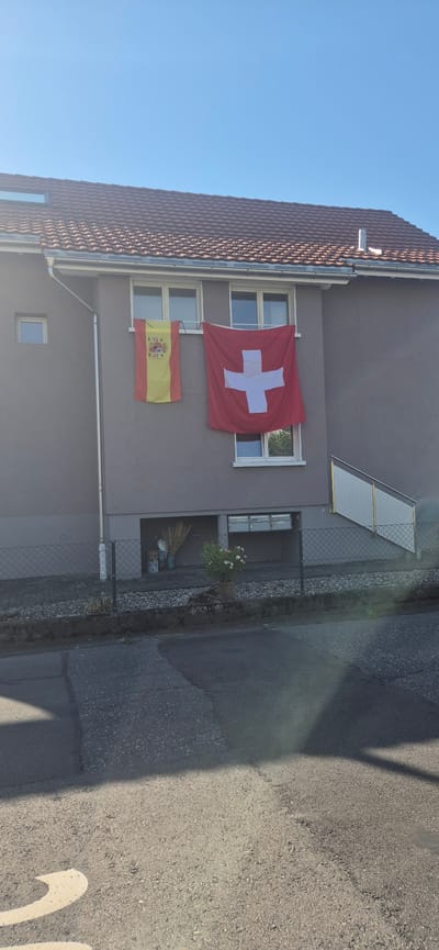 In Sargans, Switzerland, a two-story building with a dark red tiled roof is visible under a clear blue sky. Two flags are hanging from the exterior wall of the building. On the left, the flag of Spain, featuring red and yellow horizontal stripes with a coat of arms, is displayed. To its right, the flag of Switzerland, a red square with a white cross in the center, is hung. Both flags are suspended from a horizontal bar above a window. Below the flags, a lower level of the building features a row of mailboxes and a window. A metal fence encloses the area in front of this lower level. To the left of the flags, a small, square window is present on the upper floor. A white downspout runs vertically along the side of the building. The scene also shows a paved road in the foreground with shadows cast by an unseen object.