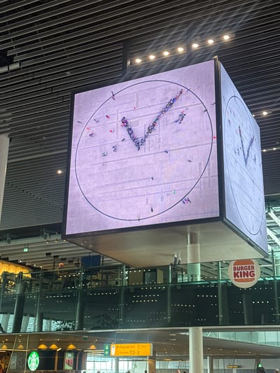The image depicts a cube-shaped video display hanging in an indoor space, likely an airport terminal, with the brand names Burger King and Starbucks visible in the background. The display shows a circular image of an overhead view of people moving about in a public space, possibly an urban plaza or open area, to form the hands of a clock, indicating time. The location is Schiphol Airport, Netherlands, as implied by the airport terminal environment. The scene includes a slatted ceiling in the foreground. A yellow sign with the words "Baggage hall" and "Transfer T2-3" is visible.