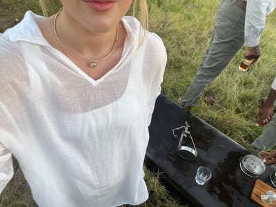 The image shows a scene in Lobilos, Botswana, featuring a partially visible person wearing a white shirt and a gold necklace standing next to a black table. Several objects are placed on the table, including a wooden board, a metal container, a glass, and a metal device with attached legs. A person standing beside the table is holding a glass and wearing grey pants and a light-colored shirt. Bare feet are visible beside the table. The surrounding environment consists of grass.