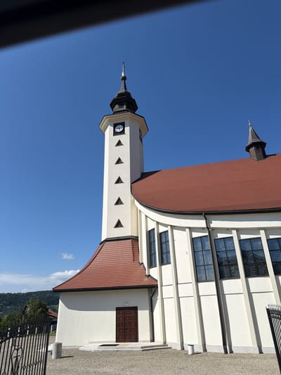 This is a vertical, eye-level photograph of a modern church with a distinct bell tower, set against a clear blue sky. The church building is predominantly white with a curved section and a red tile roof. Several windows with stained glass are visible on the main building, reflecting the sky. The bell tower, also white with a dark spire, features a clock face and a series of small, triangular windows. The scene is illuminated by bright sunlight, suggesting it is daytime, and the weather appears clear. In the background, a lush green hillside with trees and houses can be seen, hinting at a rural or semi-rural setting. A black metal fence is in the foreground on the right side of the image, with a hint of a pathway leading to the church entrance. The overall impression is one of peacefulness and architectural modernity. The photograph appears to have been taken in Mordarka, Poland. There is no visible text in the image.
