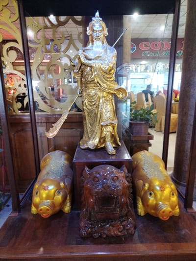 A display case containing a golden statue of a Chinese deity is flanked by two golden pig figurines and a wooden tiger figurine. The statue stands on a raised platform within the case, holding a weapon. The pig figurines are gold with reddish-pink noses. The tiger figurine is carved from wood and has an open mouth, as if roaring. The display case is made of wood with glass sides. Reflections in the glass show a restaurant interior with tables, chairs, and people. The location is in Chu Văn An Ward, Vietnam, as identified from the restaurant sign in the reflection.