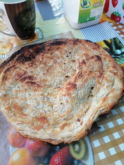 A close-up, eye-level shot focuses on a large, circular, golden-brown flatbread with visible layers and some charred areas. The bread rests on a checkered tablecloth with a fruit pattern. To the left of the bread is a white mug with brown and black accents, featuring Arabic text and the word "Coffee". Behind the mug, partially visible, is a large plastic container of white granular material, possibly sugar, with a label featuring a cartoon character and the word "MANGO". To the right of the bread, the tablecloth continues with a pattern of strawberries and other fruits. The scene appears to be outdoors due to the lighting and a hint of a bright sky in the upper right corner. There are no people visible in the image. The image is a still life of food and drink, likely on a table.
