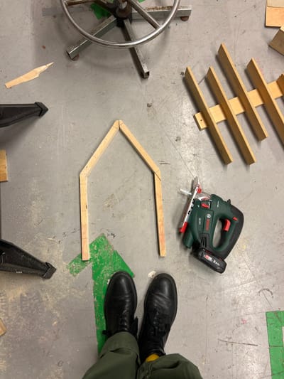 A workshop floor displays woodworking in progress. A wooden frame with angled corners lies incomplete. Nearby, a Bosch jigsaw, a wooden slatted structure, and a partially visible office chair are present. Two clamps and a small wood chip are also on the floor. A person wearing black boots and olive green pants stands adjacent to the frame. The floor is gray with a green painted arrow.