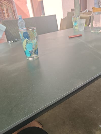 The image depicts a close-up, eye-level view of a dark gray, speckled table, likely outdoors given the background elements. On the table, to the left, is a clear drinking glass decorated with blue and green marine life motifs, such as shells and starfish. Next to it, a bottle of water is partially visible. Further to the right, a clear glass of water sits next to a glass carafe with a yellow lid, filled with a clear liquid. A small, red, cylindrical object, possibly a pen or a lighter, is placed horizontally in the center of the table. In the background, a woven chair and part of a building with a pinkish facade are visible. The overall impression is of a casual outdoor setting.