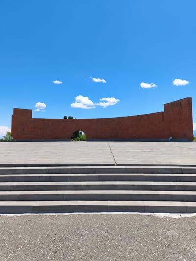 The image depicts the Tsitsernakaberd Memorial in Yerevan, Armenia, under a clear blue sky with scattered white clouds. In the foreground, a wide, paved plaza extends towards a monumental red-brick wall. This curved wall features intricate bas-relief carvings depicting historical and mythical scenes, including winged creatures and human figures. An arched opening in the center of the wall reveals glimpses of green trees and foliage beyond. Steps made of gray stone lead up to the plaza from the bottom of the frame, which shows a gravelly ground surface. The overall scene is one of solemnity and historical significance, characteristic of a memorial site.
