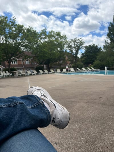 A person wearing blue jeans and white Nike Air sneakers is sitting on a concrete surface. In the background is a swimming pool with white lounge chairs, a building, and green trees under a partly cloudy sky. The location appears to be an outdoor recreational area, possibly a pool or club facility in Edison, New Jersey, United States of America.