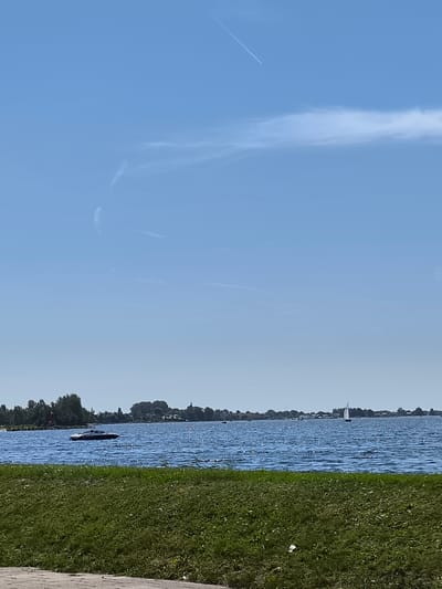 The image depicts a wide expanse of blue sky with a few wispy white clouds and a faint contrail from an aircraft. Below the sky is a body of blue water, likely a lake or canal, with a gentle ripple on its surface. A speed boat is visible on the water in the mid-left portion of the image. Further to the right, a sailboat with a white sail is also present. Along the far horizon, a tree line with buildings, including what appears to be a church steeple, suggests a settlement. The foreground consists of a strip of green grass and a paved walkway. This scene is consistent with the landscape of Aalsmeer, Netherlands, known for its waterways and proximity to nature.