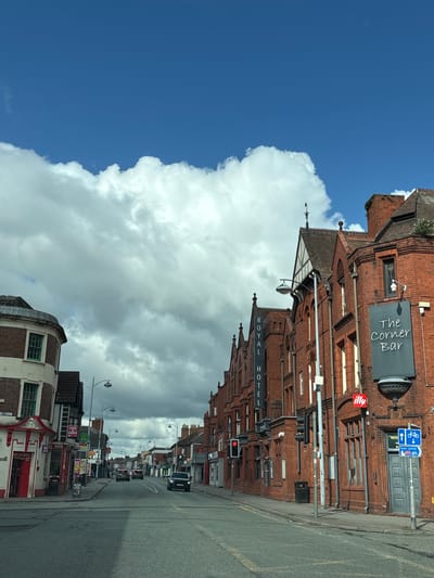The image shows a street scene in Crewe, United Kingdom. The view is of a street lined with brick buildings. "The Corner Bar" and "Royal Hotel" are identifiable establishments. A large cloud formation dominates the sky next to clear blue. Streetlights line the road, and there are traffic signals visible. Several cars are on the street. Shopfronts with signage are also visible. A street sign with "Town Centre" and a bicycle symbol is present.