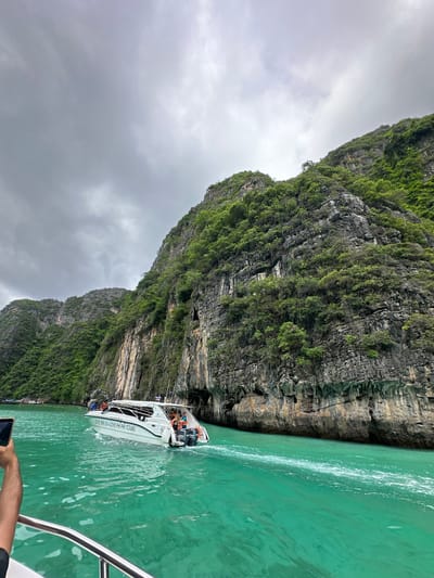The image depicts a speed boat, labeled "LOVE THE SEA PHI TOURS," navigating through vibrant turquoise water near a lush, green cliff face. The cliff is characterized by its rugged, rocky texture and dense vegetation. The sky is overcast with gray clouds. Several people are visible on the boat, some standing and others seated. In the foreground, a person's arm and hand holding a mobile phone are visible, suggesting someone is taking a photograph. The scene captures a moment of travel or tour in a scenic marine environment. The location is Haad Yao, Thailand.