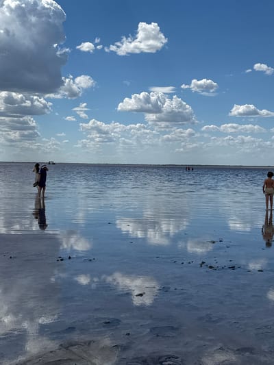 The image depicts a shallow body of water, likely a salt lake, reflecting the sky and clouds. Several individuals are visible wading in the water. On the left, two people stand close together, one appearing to be taking a photo. Further in the distance, a group of people are scattered across the water. On the right side of the frame, a single child stands facing away from the camera. The environment is open with a clear blue sky dotted with fluffy white clouds. The water is calm, with ripples distorting the reflections of the sky and the figures. The location is identified as Burbol, Russia.